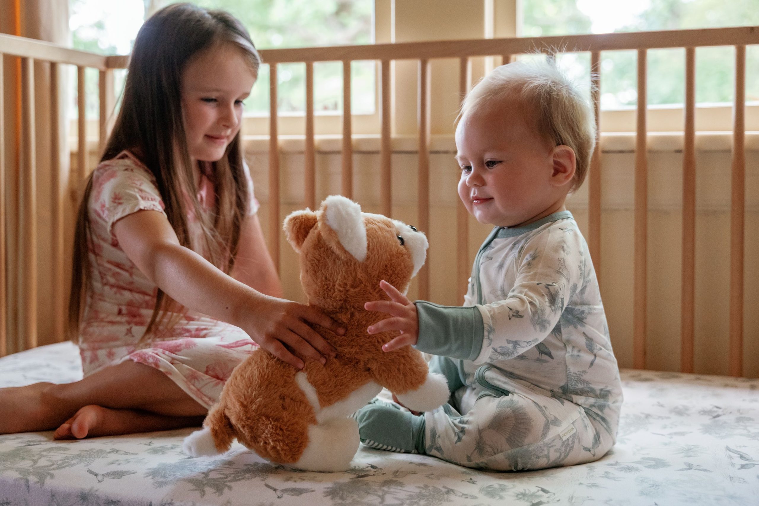 A big sister hands her baby brother a toy in the cot, both dressed in the House of Holly Birdsong print